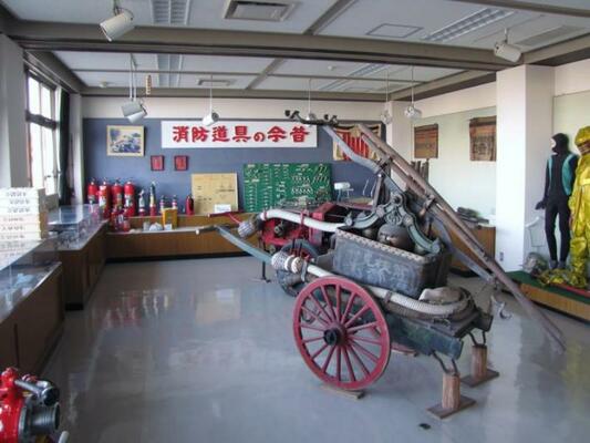 消防道具の今昔と赤字で書かれた看板の前に、消火器や大八車風の消防車など。