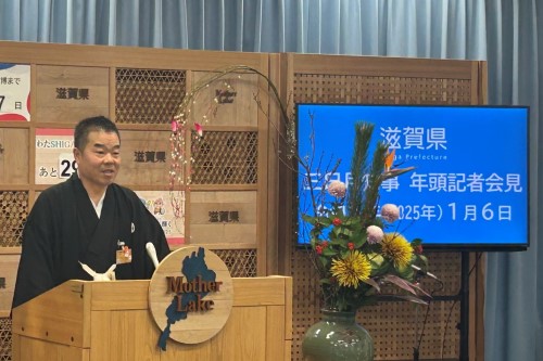 知事定例会見にて壇上で話す三日月知事の写真。知事の横に資料表示用の大きなモニターが映っている。