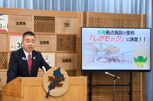 知事定例会見にて壇上で話す三日月知事の写真。知事の横に資料表示用の大きなモニターが映っている。