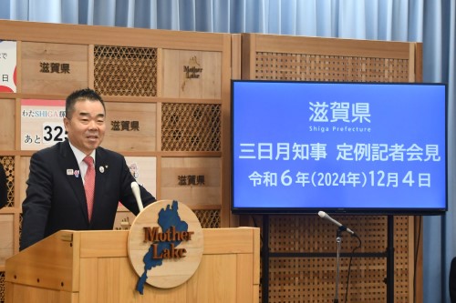 知事定例会見にて壇上で話す三日月知事の写真。知事の横に資料表示用の大きなモニターが映っている。