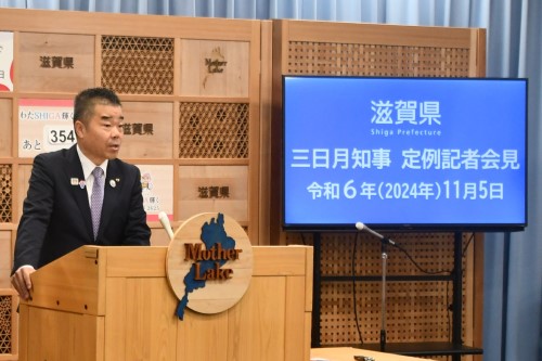 知事定例会見にて壇上で話す三日月知事の写真。知事の横に資料表示用の大きなモニターが映っている。