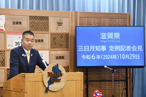 知事定例会見にて壇上で話す三日月知事の写真。知事の横に資料表示用の大きなモニターが映っている。