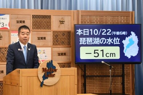 知事定例会見にて壇上で話す三日月知事の写真。知事の横に資料表示用の大きなモニターが映っている。