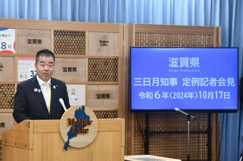 知事定例会見にて壇上で話す三日月知事の写真。知事の横に資料表示用の大きなモニターが映っている。
