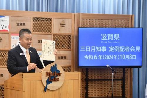 知事定例会見にて壇上で話す三日月知事の写真。知事の横に資料表示用の大きなモニターが映っている。