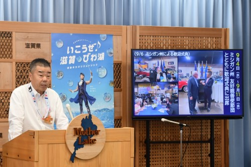 知事定例会見にて壇上で話す三日月知事の写真。知事の横に資料表示用の大きなモニターが映っている。