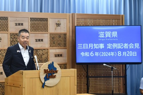 知事定例会見にて壇上で話す三日月知事の写真。知事の横に資料表示用の大きなモニターが映っている。