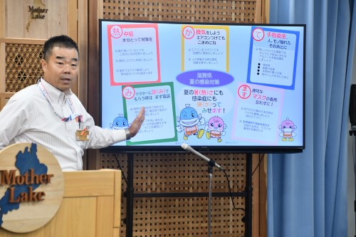知事定例会見にて壇上で話す三日月知事の写真。知事の横に資料表示用の大きなモニターが映っている。