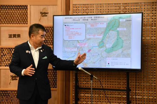 知事定例会見にて壇上で話す三日月知事の写真。知事の横に資料表示用の大きなモニターが映っている。