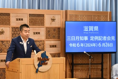 知事定例会見にて壇上で話す三日月知事の写真。知事の横に資料表示用の大きなモニターが映っている。