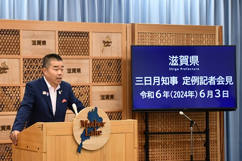 知事定例会見にて壇上で話す三日月知事の写真。知事の横に資料表示用の大きなモニターが映っている。