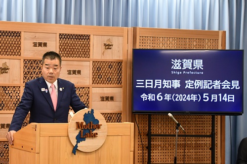知事定例会見にて壇上で話す三日月知事の写真。知事の横に資料表示用の大きなモニターが映っている。