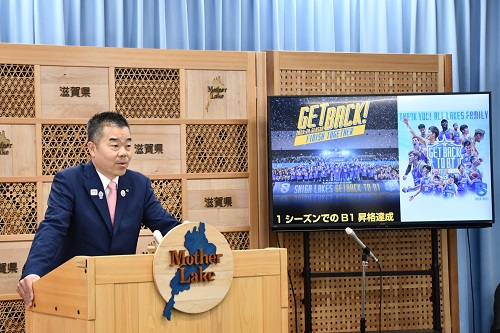 知事定例会見にて壇上で話す三日月知事の写真。プロバスケットボールのBリーグ滋賀レイクスの選手達がモニターに映っている。