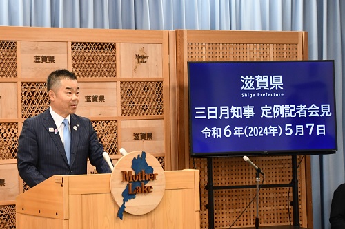知事定例会見にて壇上で話す三日月知事の写真。知事の横に資料表示用の大きなモニターが映っている。